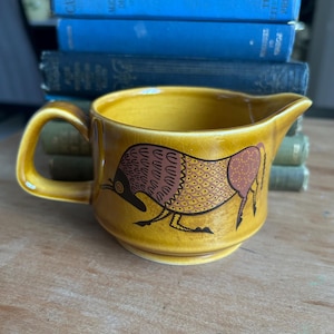 May include: A mustard yellow ceramic creamer featuring a dark brown and tan stylised animal illustration on the side. The vessel has a single handle, a pouring spout, and a visible crackle glaze finish throughout the exterior surface.