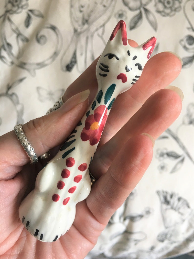 Puede incluir: Figura de gato de cer&aacute;mica blanca con detalles pintados en rojo, verde y negro. El gato tiene un cuerpo largo y delgado y un dise&ntilde;o floral en la espalda.