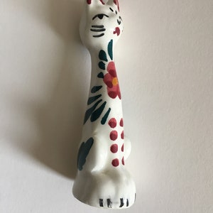 Puede incluir: Figura de cer&aacute;mica blanca de un gato con dise&ntilde;os florales pintados en verde y rojo. El gato tiene ojos y bigotes negros pintados.