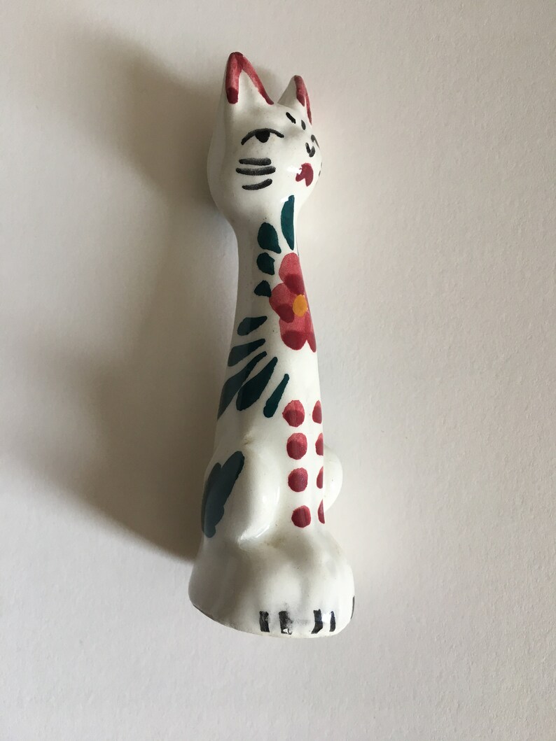 Puede incluir: Figura de cer&aacute;mica blanca de un gato con dise&ntilde;os florales pintados en verde y rojo. El gato tiene ojos y bigotes negros pintados.
