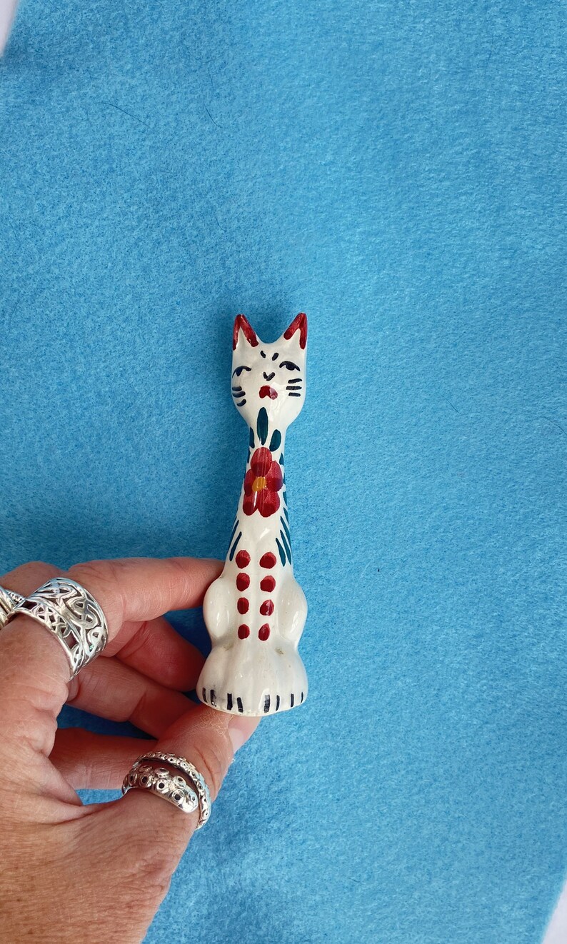 Puede incluir: Figura de cer&aacute;mica blanca de un gato con detalles pintados en rojo, azul y verde. El gato est&aacute; sentado en posici&oacute;n vertical con la cola enroscada alrededor de su cuerpo.