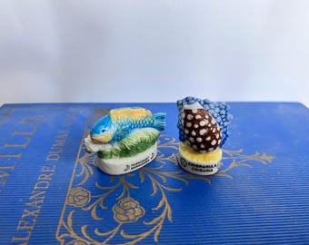 May include: Two small, colourful ceramic figurines. One is a blue and yellow fish on a green base. The other is a brown and white spotted object with blue accents. Both have text on the base.