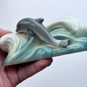 May include: A ceramic figurine of a gray dolphin leaping out of a blue and white wave. The dolphin is in mid-air with its tail and fins extended. The wave is sculpted with a swirling pattern.