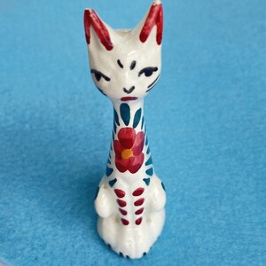 Puede incluir: Figura de cer&aacute;mica blanca de un gato con detalles pintados en rojo, azul y verde. El gato tiene un dise&ntilde;o de flor en el pecho y una cara estilizada.