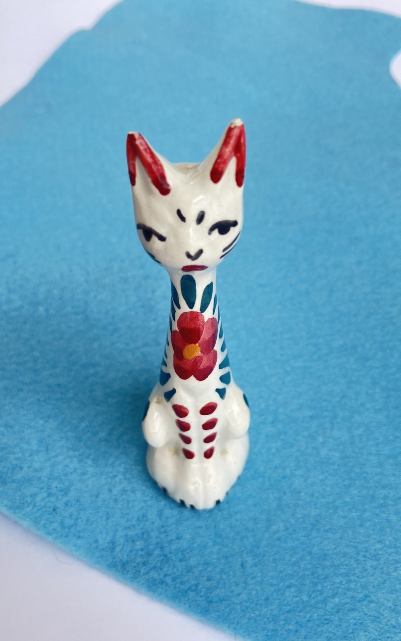 Puede incluir: Figura de cer&aacute;mica blanca de un gato con detalles pintados en rojo, azul y verde. El gato tiene un dise&ntilde;o de flor en el pecho y una cara estilizada.