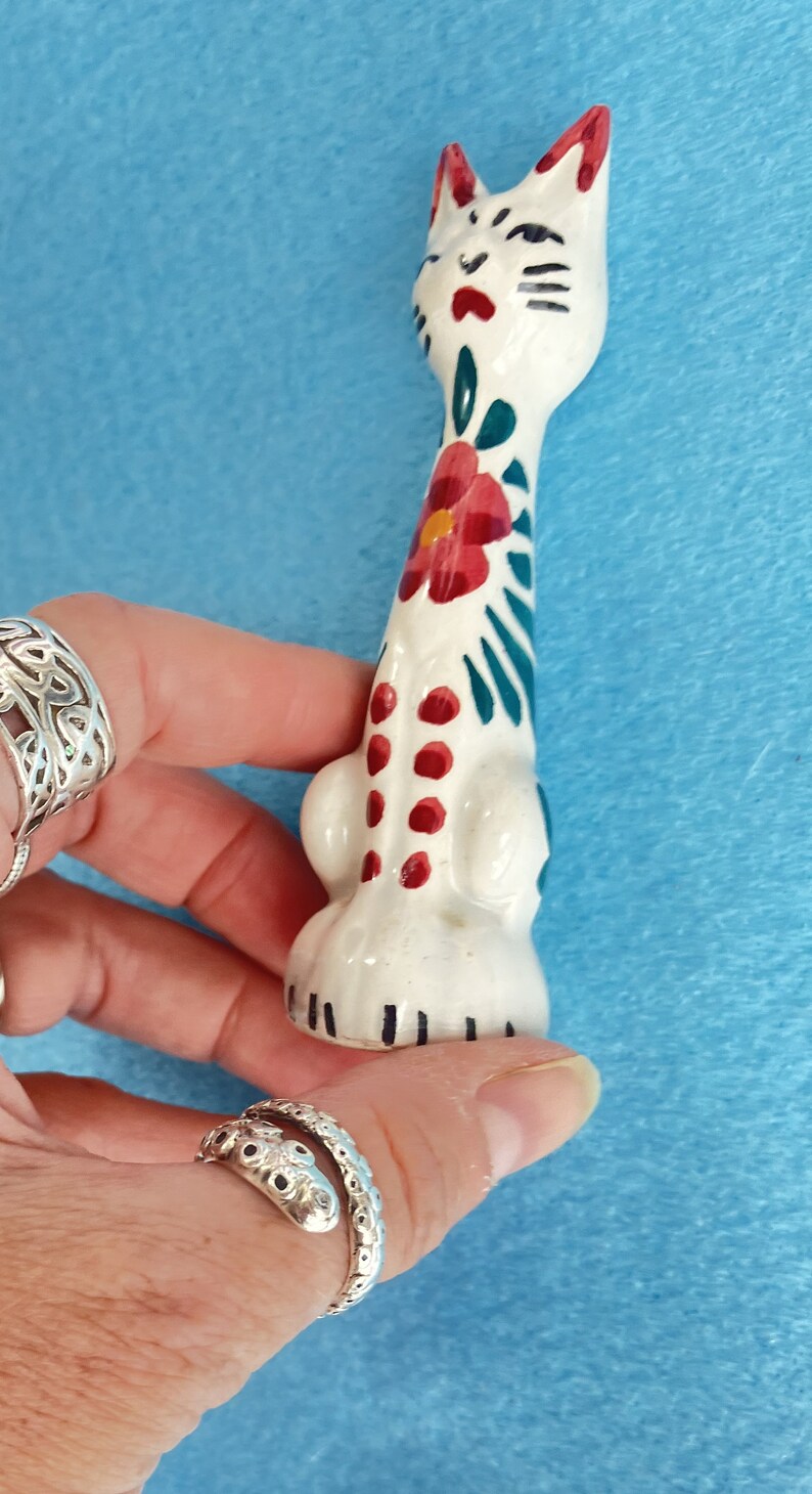 Puede incluir: Figura de cer&aacute;mica blanca de un gato con dise&ntilde;os florales verdes y rojos. El gato est&aacute; de pie con la cabeza inclinada hacia arriba.
