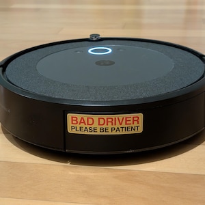 May include: A black round robot vacuum cleaner with a gold sticker that says "Bad Driver Please Be Patient".