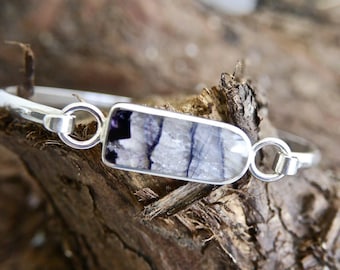 May include: A silver bracelet with a rectangular, bezel-set stone. The stone features a mix of white, gray, and purple hues. The bracelet is resting on a piece of textured, brown wood.