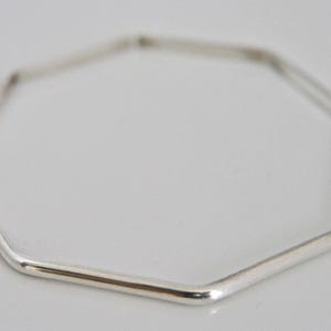 May include: A silver-coloured, geometric bangle bracelet. The bracelet has a polygonal shape with smooth, rounded edges. The bangle is made of a shiny, reflective metal, and is displayed against a plain white background. The bracelet is a piece of jewellery.
