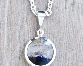 May include: A silver pendant necklace featuring a round, polished stone with a mix of colors. The stone has a translucent appearance with shades of white, gray, and deep blue. The pendant hangs from a silver chain.