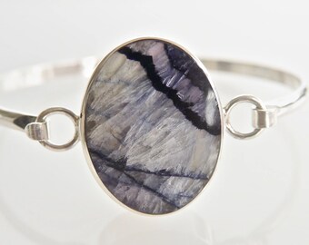 May include: A silver bracelet featuring an oval-shaped gemstone with a unique pattern of blue, gray, and black hues. The gemstone is set in a silver bezel, and the bracelet has a simple, elegant design.