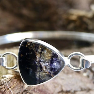 May include: A silver bangle bracelet featuring a triangular dark stone with a mix of black, brown, and purple tones. The bracelet is displayed against a blurred wooden backdrop, emphasizing the jewellery's design.