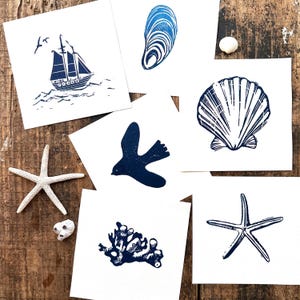 May include: A selection of navy blue and white art prints with a nautical theme, including a sailboat, seashell, starfish, bird, mussel, and coral. The prints are displayed on a wooden surface, creating a coastal atmosphere.