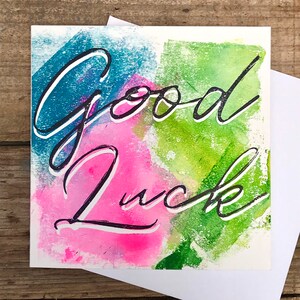 May include: A watercolour card with the words "Good Luck" written in black cursive script. The background is a mix of pink, blue and green watercolour washes.