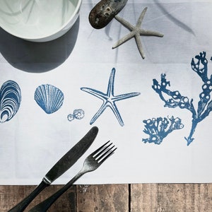 May include: A white linen table runner with blue printed designs of seashells, starfish, and seaweed. Two silver utensils are placed on the table runner.