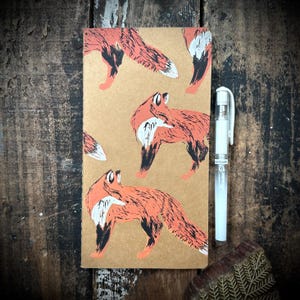 May include: A brown notebook with a repeating pattern of orange foxes printed on the cover. The foxes are in various poses, some are facing forward, some are facing sideways, and some are facing backward. The notebook is closed and has a white pen lying next to it.