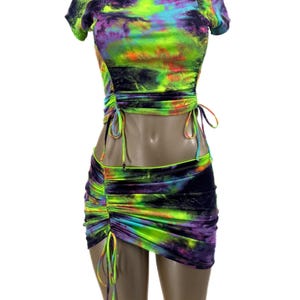 May include: A two-piece outfit with a tie-dye print in shades of green, purple, and black. The top is a cropped, short-sleeved shirt with a drawstring at the waist. The skirt is a mini skirt with a drawstring at the waist.
