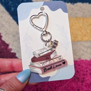 May include: A keychain featuring a stack of books with the words "Book Lover" and a heart. The keychain has a silver heart-shaped clasp and is attached to a light blue card with a floral design.