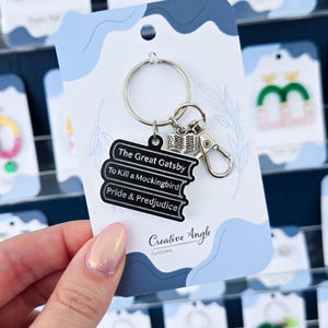 May include: A silver keychain with a stack of three black books, each labeled with a classic novel title: "The Great Gatsby," "To Kill a Mockingbird," and "Pride & Prejudice." The keychain also features a small open book charm and a lobster clasp.