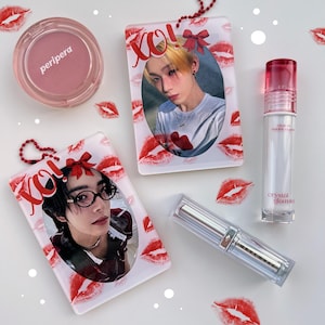 Könnte beinhalten: Draufsicht auf Schönheitsprodukte und Accessoires. Enthält einen rosa Rouge-Kompaktpuder, einen transparenten Lipgloss und eine silberne Lippenstift-Tube. Zwei transparente rechteckige Schlüsselanhänger mit Fotos und roten Lippenabdrücken sind ebenfalls vorhanden.