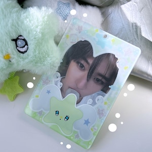 May include: A light green star-shaped plush toy and a photo card holder with a picture of a person. The photo card holder has a star and cloud design with a light green and blue colour scheme. The plush toy is light green and fluffy.