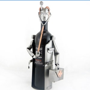 Puede incluir: Un porta botellas de vino de metal plateado con forma de médico con un estetoscopio y un botiquín de primeros auxilios. El porta botellas está sujetando una botella de vino.