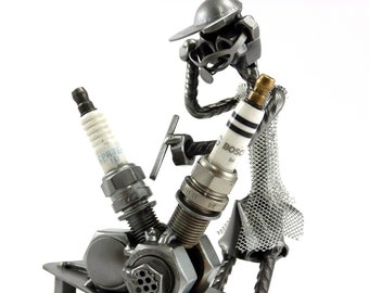 Può includere: Una scultura in metallo di una persona che lavora su un piccolo motore. La persona è fatta di metallo e indossa un cappello e degli occhiali. Il motore è fatto di metallo e ha candele e una chiave inglese. La scultura è su un piccolo tavolo di metallo.