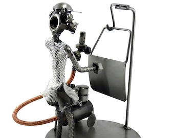 Può includere: Una scultura in metallo di una persona che tiene una chiave inglese e seduta su un compressore d'aria. La scultura è realizzata con varie parti metalliche, tra cui dadi, bulloni e rondelle. La persona indossa un casco e occhiali di sicurezza. La scultura è montata su una base in metallo.