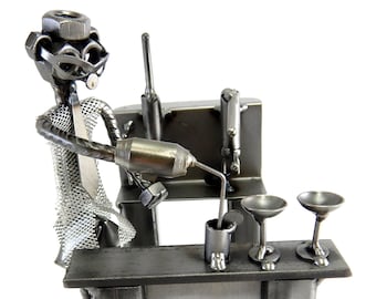 May include: A metal sculpture of a bartender pouring a drink at a bar. The sculpture is made of nuts, bolts, and other metal scraps. The bartender is wearing a tie and has a pair of glasses. The bar has two glasses on it.