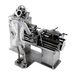 Könnte beinhalten: Eine Metallsculptur einer Person, die eine Drehbank bedient. Die Skulptur besteht aus verschiedenen Metallteilen, darunter Muttern, Schrauben und Zahnrädern. Die Person trägt ein Netzhemd und einen Hut. Die Drehbank ist ein kleines Tischmodell mit einem Metallbett und einer Spannzange.