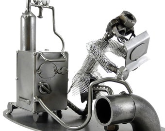 May include: A metal sculpture of a person sitting on a pipe, holding a laptop. The person is made of nuts, bolts, and other metal pieces. The sculpture is on a metal base.