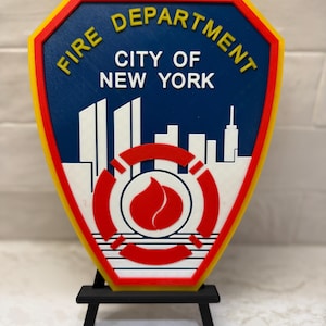 May include: A 3D-printed Fire Department badge from New York City, featuring a shield shape with a yellow border, red trim, and a blue center. The badge displays the text "FIRE DEPARTMENT CITY OF NEW YORK" in yellow and white, with a skyline and a red Maltese cross.