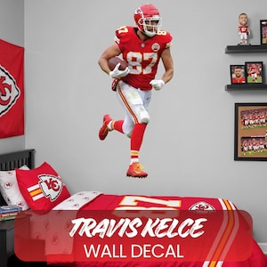 Op de afbeelding: Een muursticker met Travis Kelce in een rood en wit voetbaluniform, rennend met een bal. De sticker is geplaatst boven een bed met bijpassend rood en wit beddengoed. De tekst "TRAVIS KELCE WALL DECAL" is zichtbaar.