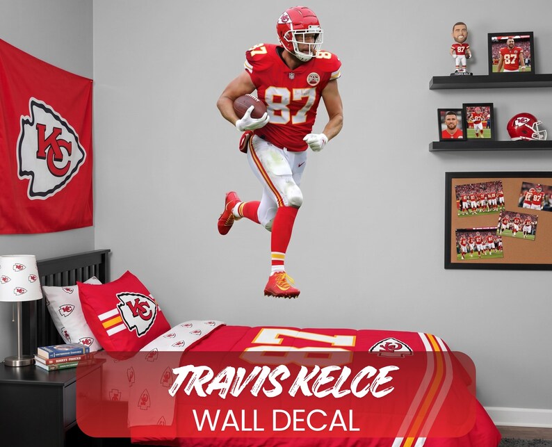 Op de afbeelding: Een muursticker met Travis Kelce in een rood en wit voetbaluniform, rennend met een bal. De sticker is geplaatst boven een bed met bijpassend rood en wit beddengoed. De tekst "TRAVIS KELCE WALL DECAL" is zichtbaar.