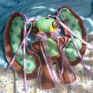 May include: A handmade fabric doll featuring brown and mint green polka dot fabric with visible pink blanket stitching. The doll has large ears, button eyes, and a decorative light purple ribbon tied in a bow at the center of its chest.