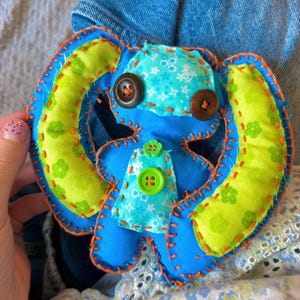 May include: A handmade blue plush bunny with orange stitching, green and blue patterned accents, and brown button eyes. The bunny has large ears and a small body, and is made of soft fabric.