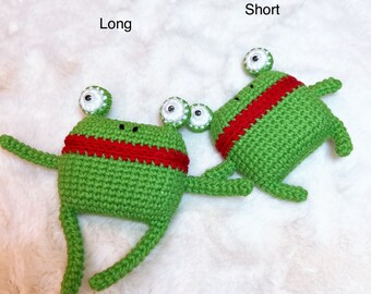 May include: Two green crocheted frog plushies with red and white accents. The frog on the left has longer legs and is labeled "Long". The frog on the right has shorter legs and is labeled "Short".