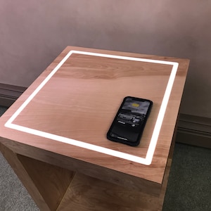 May include: A wooden side table with a built-in illuminated square border. A smartphone with a black case rests on the table, displaying a car image. The table has a natural wood finish and a simple, modern design.