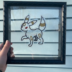 May include: A framed artwork featuring a cartoon character with a white body, gold accents, and red eyes. The character is set against a clear background within a black frame. The frame is held by a hand.