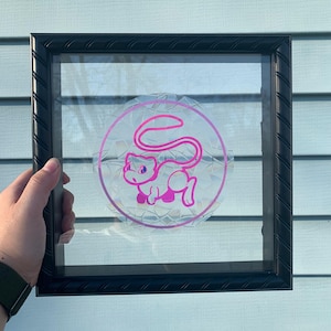 May include: Black framed art featuring a pink cartoon character inside a circle. The character has a long tail and is set against a clear, textured background. The frame is square with a decorative edge.