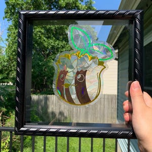 May include: A black, twisted-frame shadow box containing a holographic sticker of a stylized, anthropomorphic fruit character with brown vertical stripes, large white eyes, and a green leaf outline, featuring a mosaic-like reflective surface.