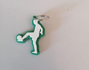 Op de afbeelding: Een groen en witte sleutelhanger met een voetballer die een bal trapt. De sleutelhanger heeft een zilveren metalen ring om aan sleutels of een tas te bevestigen. De voetballer is wit, met een groene omtrek.