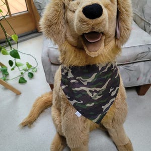 May include: A plush golden retriever dog with a camouflage bandana. The dog is sitting on a light-colored surface, with a patterned chair in the background. The dog has a black nose and open mouth.