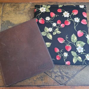May include: A dark brown leather e-reader case with the word amazon embossed on the bottom right corner, paired with a rectangular fabric pouch featuring a black background with a repeating pattern of red strawberries, green leaves, and white flowers.