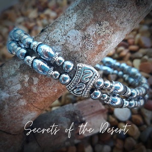 May include: A silver bracelet with a double row of beads and a decorative silver charm. The bracelet is on a wooden surface with the text "Secrets of the Desert" below it.