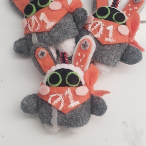 May include: Three handmade felt plush keychains featuring a grey body, orange ears with grey cross-stitch details, and a bright orange bandana with the white embroidered number 01. Each character has large black eyes with green outlines and pink cheeks.