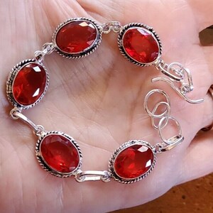May include: A silver bracelet with five red oval-shaped gemstones. The bracelet has a lobster claw clasp.