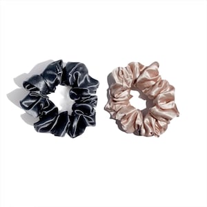 May include: Two satin scrunchies, one in a dark charcoal gray and the other in a light peach color, are displayed against a white background. The scrunchies are round and gathered, with a shiny, smooth texture.