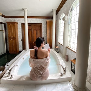 May include: A person sits in a white jacuzzi tub, reading a book. The person is wearing a light pink shirt. The bathroom has white columns, a large window, and wooden cabinets. A glass of wine sits on a tray.