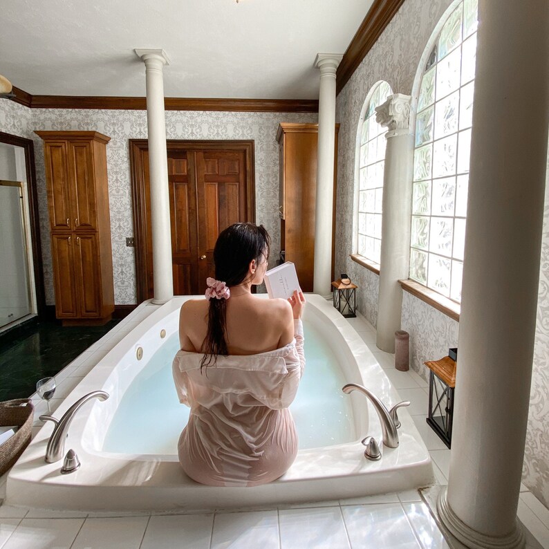 May include: A person sits in a white jacuzzi tub, reading a book. The person is wearing a light pink shirt. The bathroom has white columns, a large window, and wooden cabinets. A glass of wine sits on a tray.